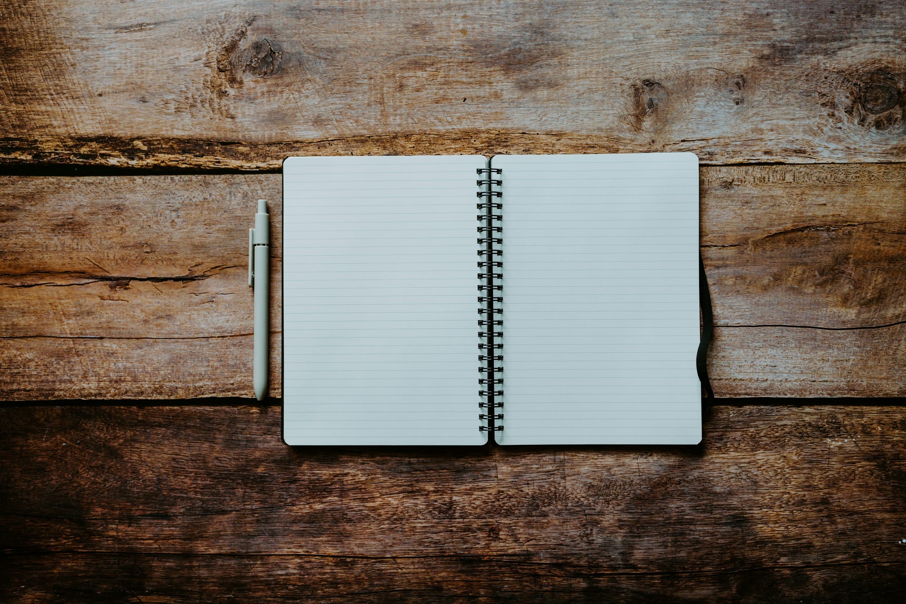 An open lined spiral notebook and a pale pen on a rough wooden table.