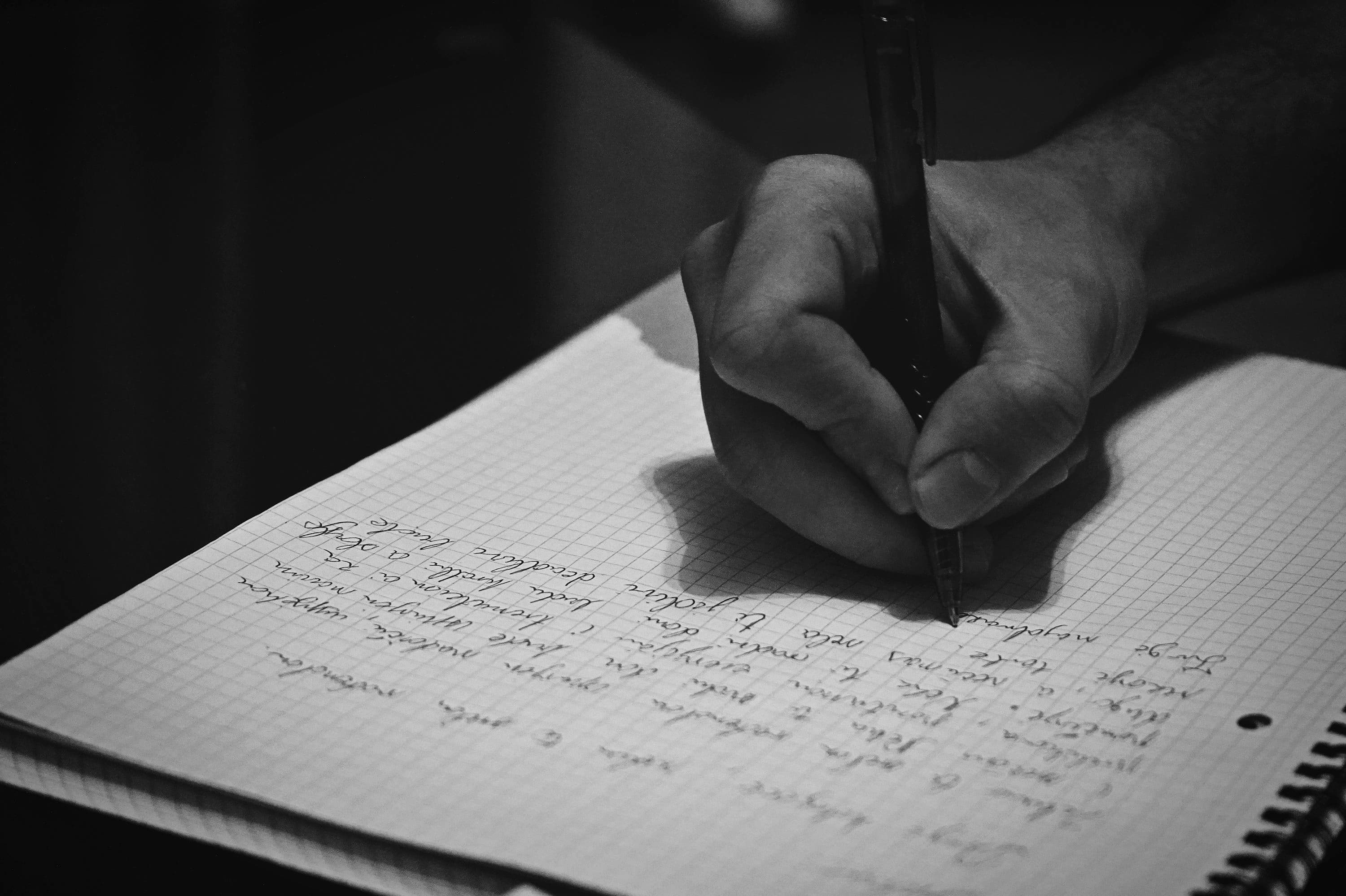 A black-and-white close-up of a hand writing with a pen in a squared-paper notebook.