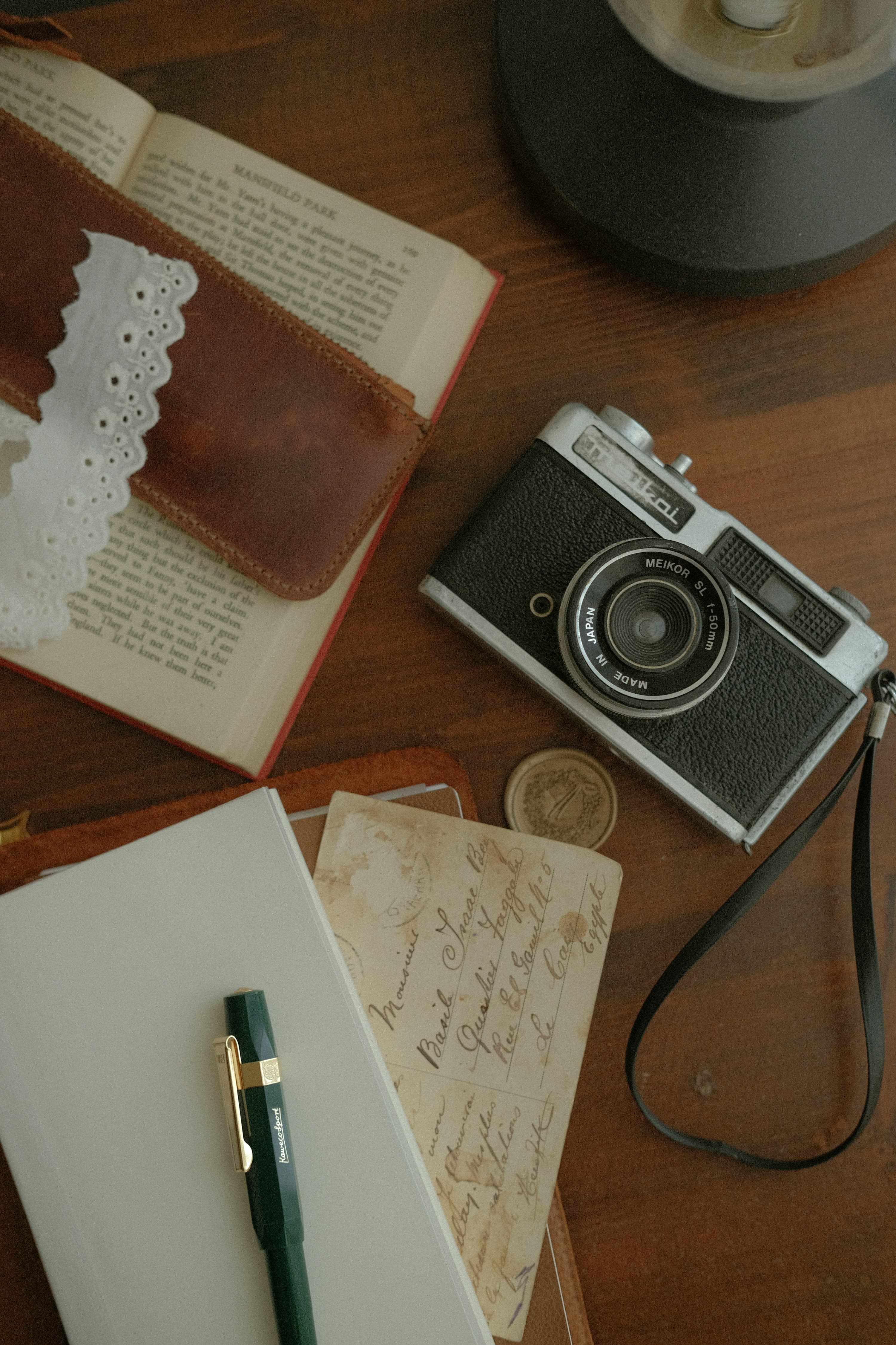 An open notebook and green fountain pen beside an old handwritten postcard, a book and a vintage camera on a wooden table.